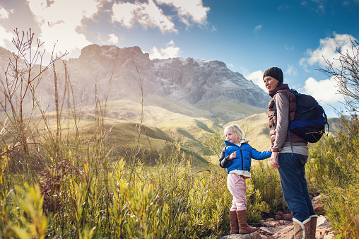 Mother with backpack and young blonde girl holding hands in nature with mountains with snow and plants Jonkershoek Stellenbosch Cape Town South Africa