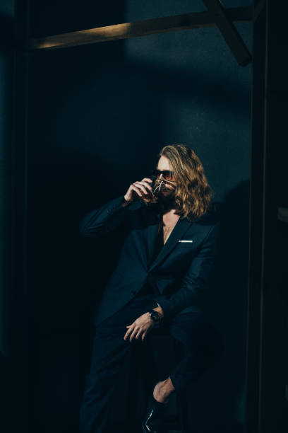istock Handsome stylish long haired man in suit drinking whiskey on black 817513632