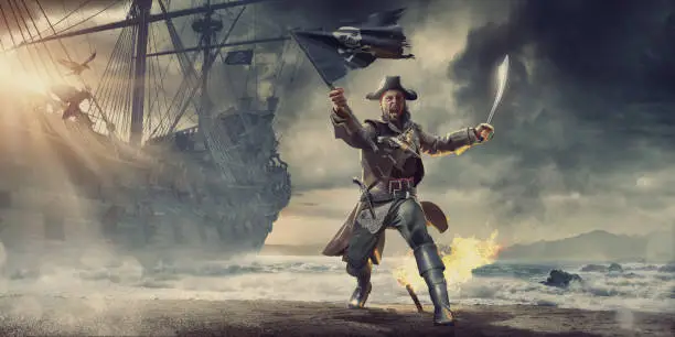 A conceptual image of male pirate on a beach behind a pirate ship on an stormy overcast evening. The pirate is holding a torn pirate flag and cutlass with mouth open shouting. He has a flaming torch in the sand behind him, and wears pirate clothes and has guns in holsters.