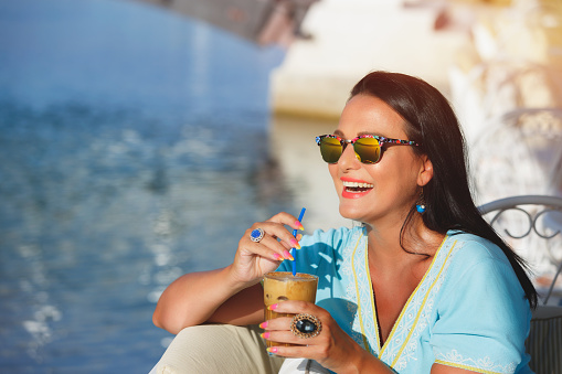 Happy woman wearing sunglasses, holding glass of iced coffee and looking away smiling, selective focus, vintage toned image
