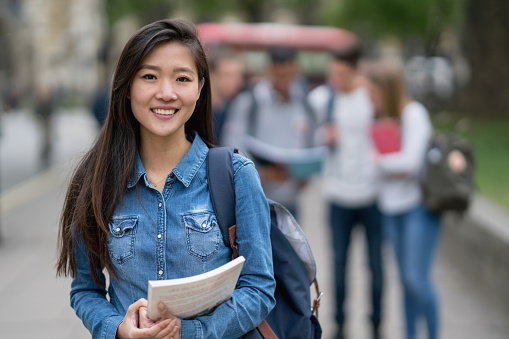 Portrait of an Asian student on the street looking at the camera smiling with a group at the background - abroad education concepts