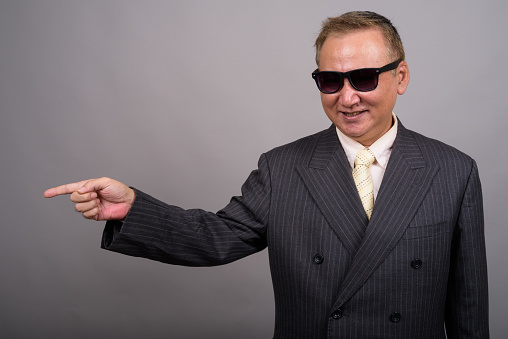 Studio shot of mature Asian businessman wearing sunglasses looking cool against gray background horizontal shot