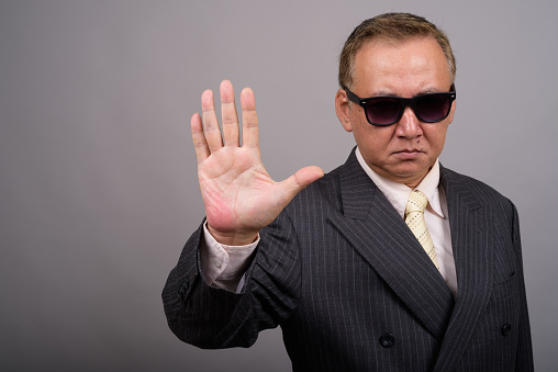 Studio shot of mature Asian businessman wearing sunglasses looking cool against gray background horizontal shot
