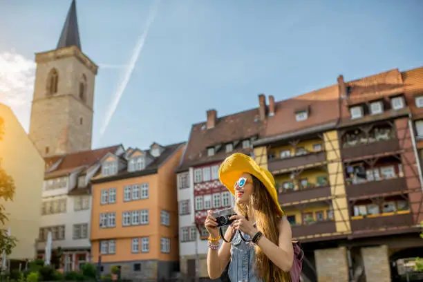 Young woman tourist in yellow hat standing with photo camera on the famous Merchants bridge background in Erfurt city, Germany