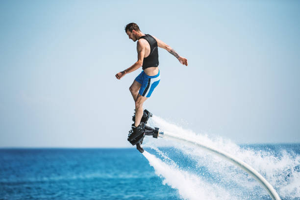 Flyboarding. Closeup side view of a young man enjoying flyboarding session at sea on a sunny summer day. He's wearing blue shorts and black life jacket. hoverboard stock pictures, royalty-free photos & images