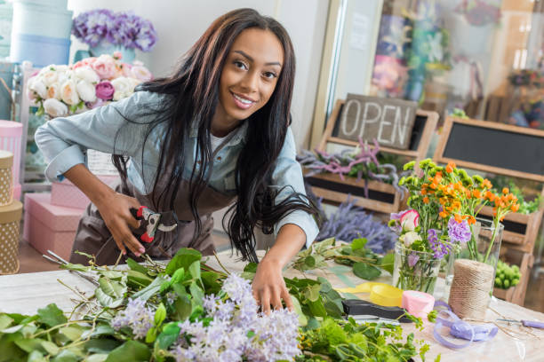 beautiful african american woman in apron holding secateurs while cutting flowers and smiling at camera in flower shop beautiful african american woman in apron holding secateurs while cutting flowers and smiling at camera in flower shop the black womens expo stock pictures, royalty-free photos & images