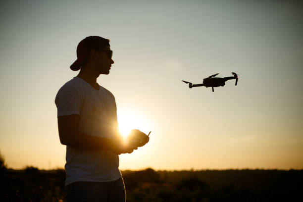 istock Silhouette of a man piloting drone in the air with a remote controller in his hands on sunset. Pilot takes aerial photos and videos with quadcopter from above 818399282
