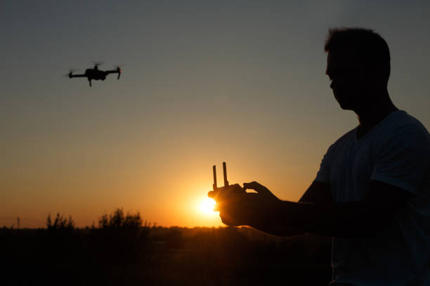 istock Silhouette of a man piloting drone in the air with a remote controller in his hands on sunset. Pilot takes aerial photos and videos with quadcopter from above 818401878