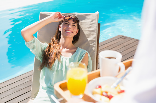 Smiling young woman looking at cropped waiter with breakfast tray by the pool