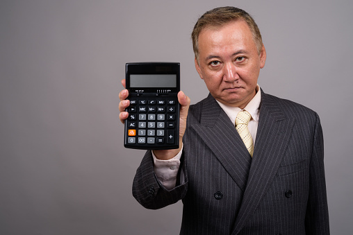 Studio shot of mature Asian businessman holding calculator against gray background horizontal shot