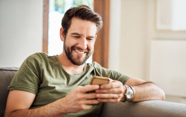 Let’s see what everyone’s doing this weekend Shot of a happy young man using a mobile phone on the sofa at home bachelor stock pictures, royalty-free photos & images