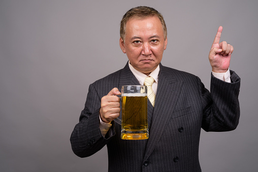 Studio shot of mature Asian businessman holding glass of beer against gray background horizontal shot