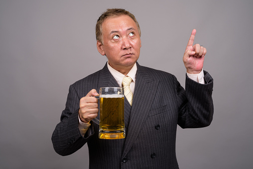 Studio shot of mature Asian businessman holding glass of beer against gray background horizontal shot
