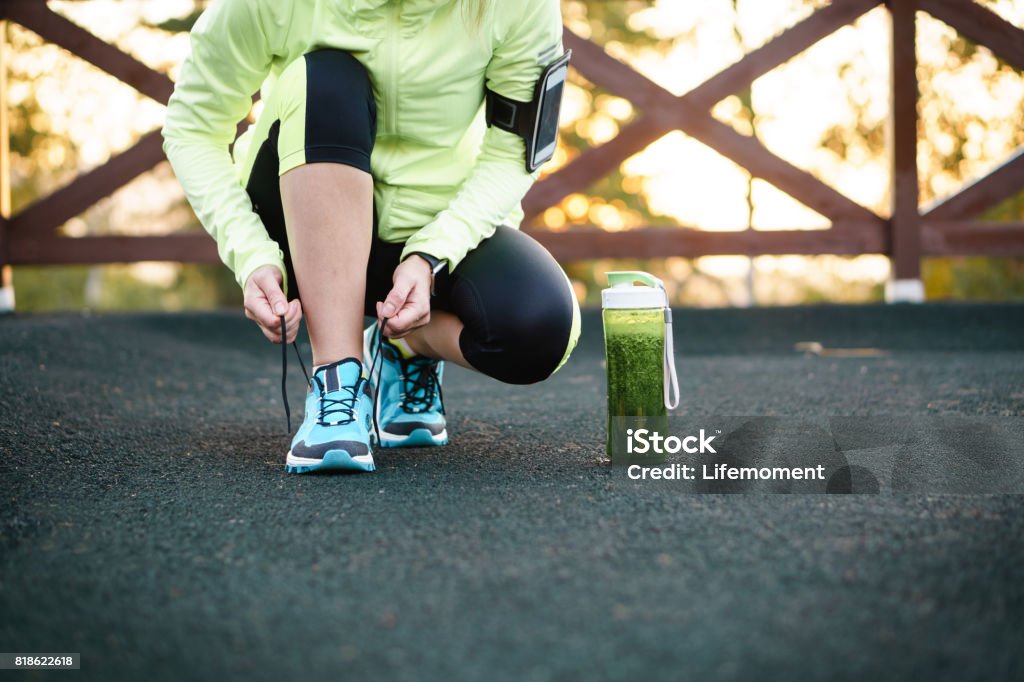 Green detox smoothie cup and woman lacing running shoes before workout. Green detox smoothie cup and woman lacing running shoes before workout. Fitness and healthy lifestyle concept. Exercising Stock Photo