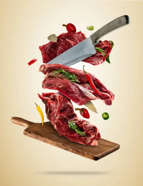 Flying pieces of raw steaks, with ingredients for cooking, served on woodenboard. Knife cuting the meat. Concept of food preparation in low gravity mode. Separated on smooth background. High resolution image