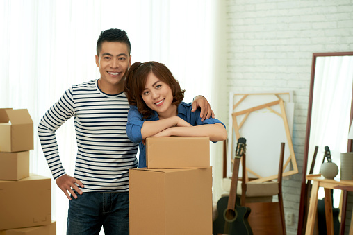 Group portrait of handsome young man gently embracing his girlfriend while she leaning on cardboard box and looking at camera with charming smile, they are ready to move into new apartment