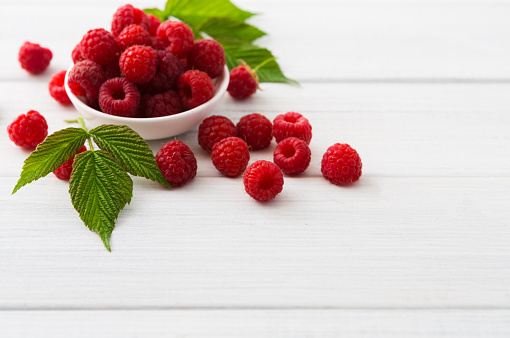 Red fresh raspberries on white rustic wood background. Bowl with natural ripe organic berries with peduncles and green leaves on wooden table, top view with copy space