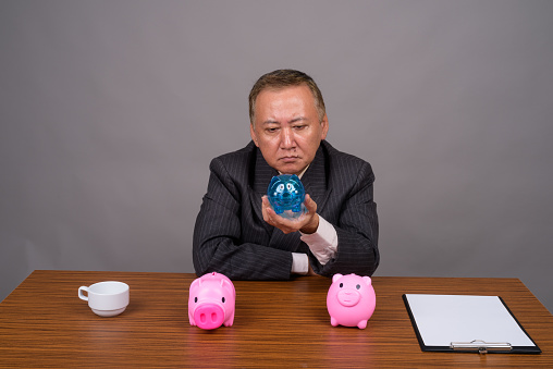 Studio shot of mature Asian businessman sitting with wooden table against gray background horizontal shot