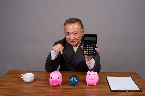 Studio shot of mature Asian businessman sitting with wooden table against gray background horizontal shot