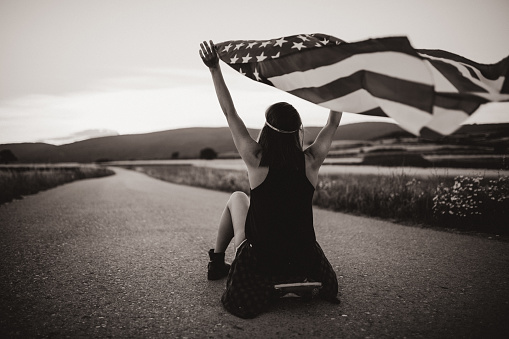 One woman, riding her skateboard on the street far from the city, in nature holding US flag, rear view, black and white.