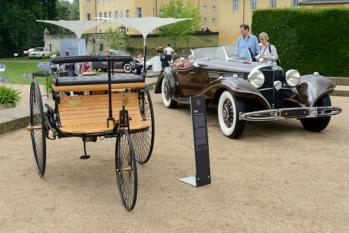 Benz Patent Motor-wagen 1886 the world's first automobile - a vehicle designed to be propelled by an internal combustion engine with a Mercedes-Benz 500K Luxus Roadster 1930s luxury convertible touring car in the background. The cars are on display during the 2016 Classic Days event at Schloss Dyck. People in the background are looking at the cars.