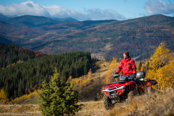 homme en veste et chapeau à cheval sur un vtt rouge sur les routes de montagne sur une journée ensoleillée d’automne. beau paysage de forêts, de montagnes et de bleu ciel avec espace copie - off road vehicle quadbike quad racing motocross photos et images de collection