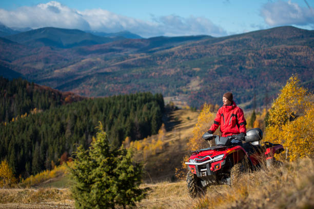 man in jacket and hat riding on a red atv on mountain roads on a sunny autumn day. beautiful landscape of forest, mountains and blue sky with copy space - off road vehicle snow 4x4 driving imagens e fotografias de stock