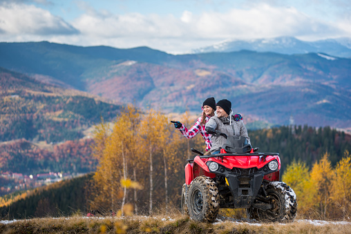 Happy couple on red quad bike riding in the mountains. Young man driving the ATV, girlfriend pointing at something interesting. Beautiful landscape of mountains and forest at autumn day