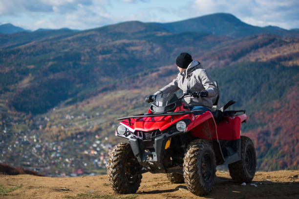 hombre en ropa de invierno en una cuatrimoto roja en la cima de la montaña disfrutando de las vistas de las montañas poderosas detrás de él. fondo borroso del paisaje de montaña - off road vehicle snow 4x4 driving fotografías e imágenes de stock