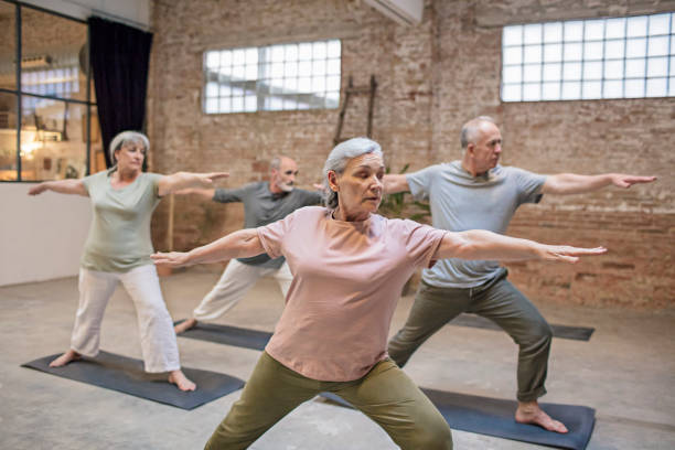 Senior people standing in warrior pose at gym Active senior woman with friends standing in warrior pose at gym. Males and females are exercising in health club. They are wearing sports clothing at yoga studio. warrior 2 stock pictures, royalty-free photos & images