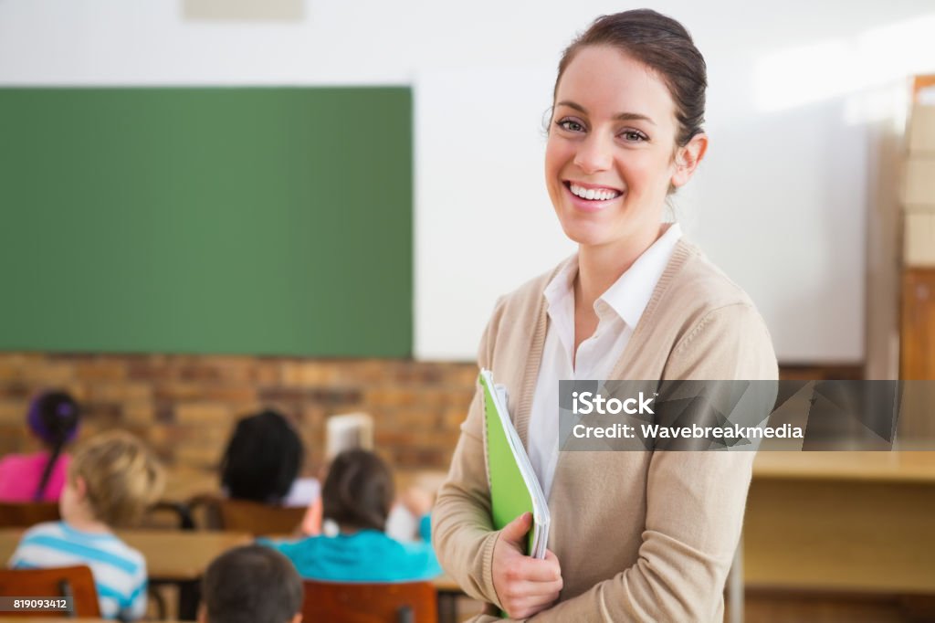 Professora bonita sorrindo para a câmera na parte traseira da sala de aula - Foto de stock de Professor royalty-free