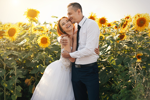 Bride redhead in wedding white dresses and groom in whit shirt and tie standing hugging in the field of sunflowers. With sunshine summer