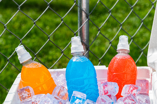 A red and white ice chest full of ice and several plastic bottles of colorful ice cold sports drinks on a hot Summer day with a chain link fence in the background.