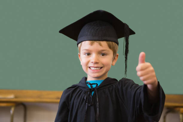 Cute pupil in graduation robe smiling at camera in classroom at the elementary school