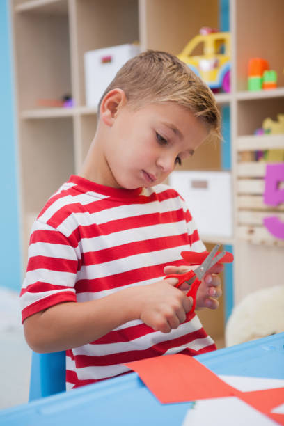 istock Cute little boy cutting paper shapes in classroom 819424756
