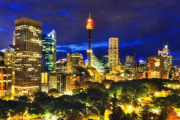 Sy Hyde Park Towers Dark Panorama of Sydney city high-rise towers illuminated at sunset standing over green trees of the central Hyde Park. hyde park sydney stock pictures, royalty-free photos & images
