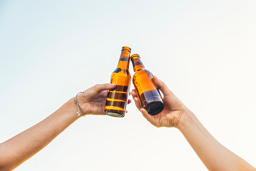 Female friends cheers clinking bottles of beer in hands in the park.