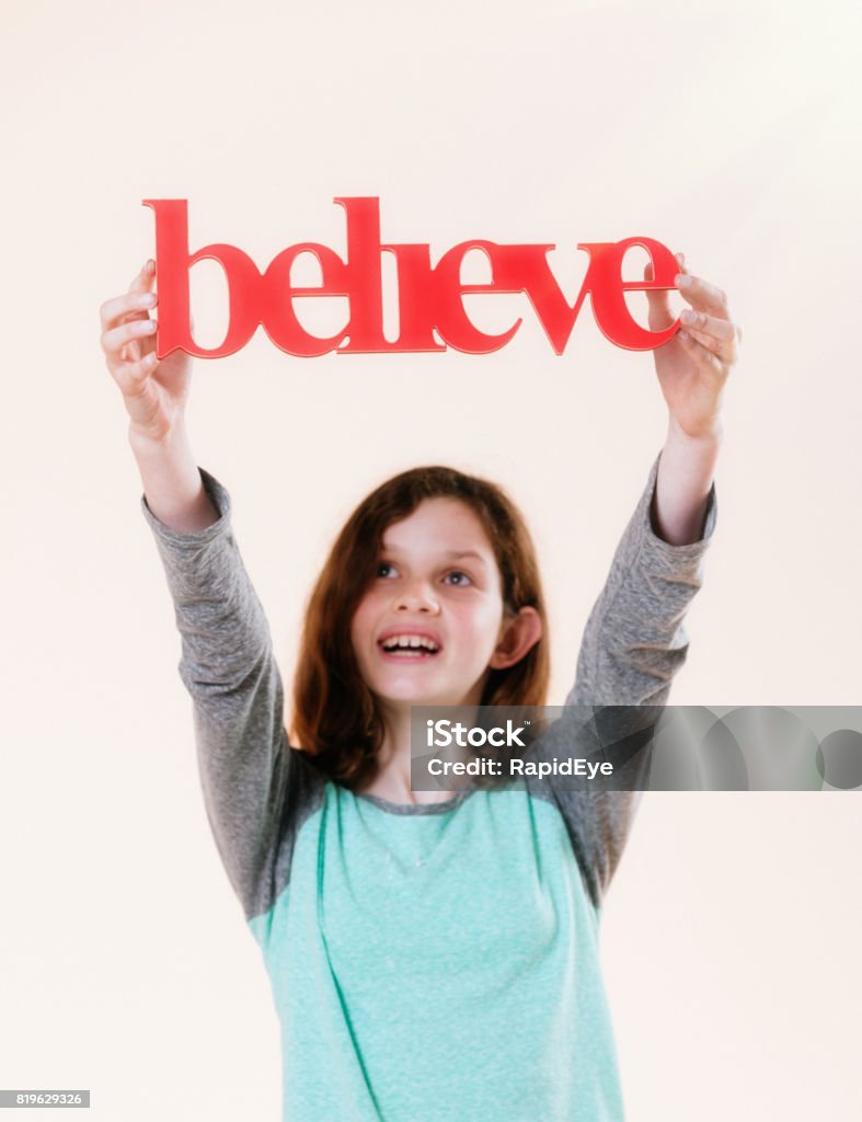 'believe' says three-dimensional sign held aloft by smiling pre-teen girl A pretty 12-year-old girl smiles as she holds up a 3D sign reading "believe". Child Stock Photo