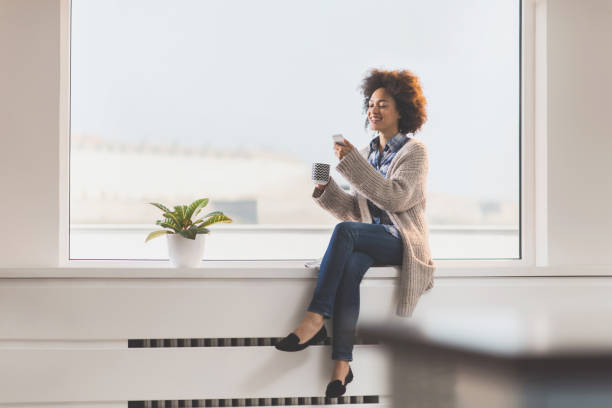 istock Beautiful African American woman using mobile phone while relaxing on a window sill. 819636790