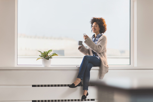 Happy black woman sitting on window sill with cup of coffee and typing text message on mobile phone. Copy space.