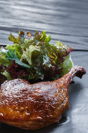 Roasted duck leg closeup, served on slate plate with lettuce. Restaurant food on black wood table