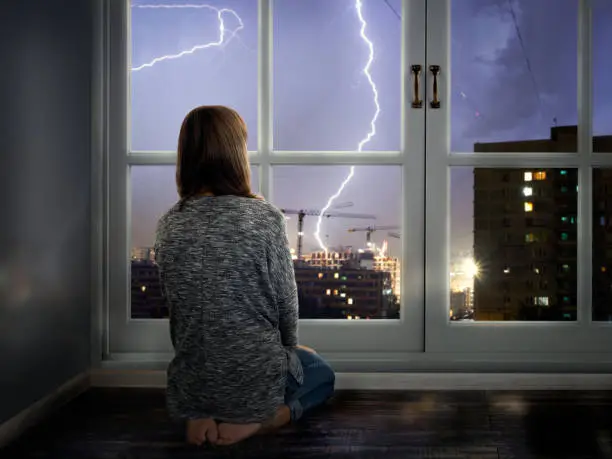 The girl looks through the window at the lightning. Thunderstorm in the city