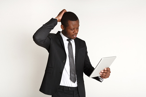 Pensive businessman using tablet isolated on white. Young african american man in suit has problem with device or read bad news, touching his head
