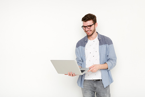 Young man standing with laptop. Handsome guy in glasses holding open notebook in his hand and work on it. Information, technology concept