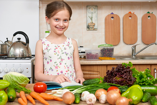 child girl with fruits and vegetables in home kitchen interior, read cooking book, healthy food concept