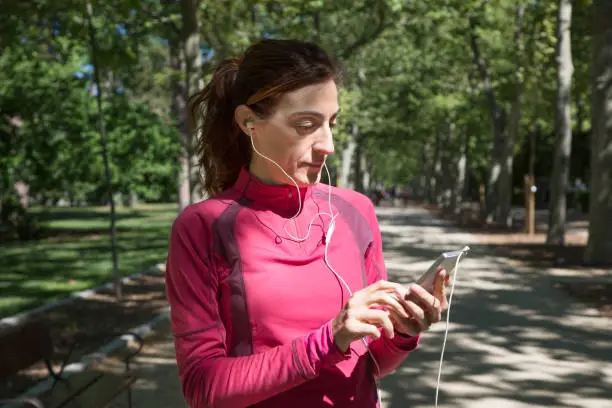 sporty adult woman with red sweater, with earphones watching mobile smartphone in park of Retiro, in Madrid, Spain. Horizontal shot