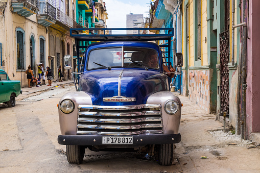 A man is sitting inside an old american truck of the 50s in Havana city center. Other people in the background are talking
