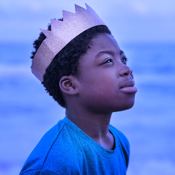 Portrait of a young african-american boy on the beach at dusk. He has a serious face, wearing a gold paper crown. He is wearing a blue t-shirt and the image has purple and blue tone. Square head and shoulders outdoors shot with copy space. This was taken in Florida, USA.