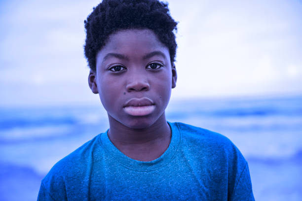 Portrait of a young african-american boy on the beach at dusk. He has a serious face, looking at the camera. He is wearing a blue t-shirt and the image has purple and blue tone. Horizontal head and shoulders outdoors shot with copy space. This was taken in Florida, USA.