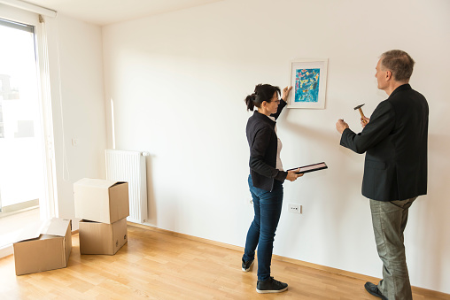 Mature adult couple in new, very bright empty apartment.  They stand on parquet floor and planing where to hang a painting on white wall. They stand at large, glass door to the balcony, in background cardboard boxes.